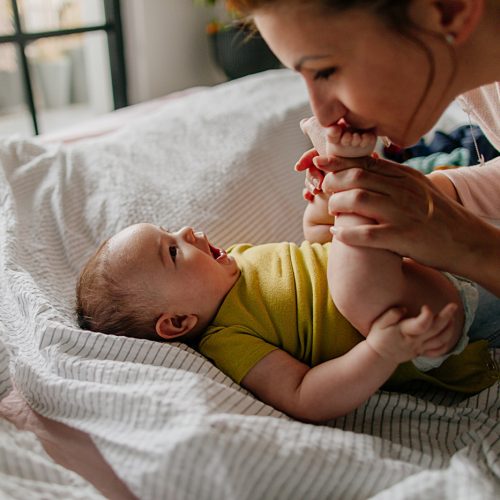 Portrait of a little smiling baby boy and his mom, kissing his tiny feet right after waking up in his nursery