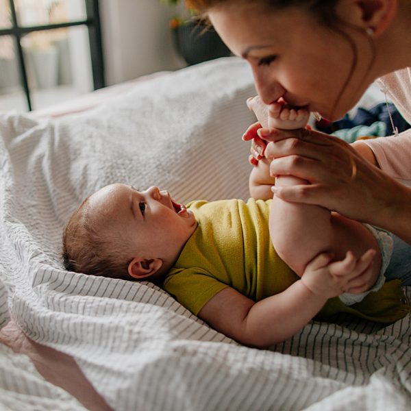 Portrait of a little smiling baby boy and his mom, kissing his tiny feet right after waking up in his nursery