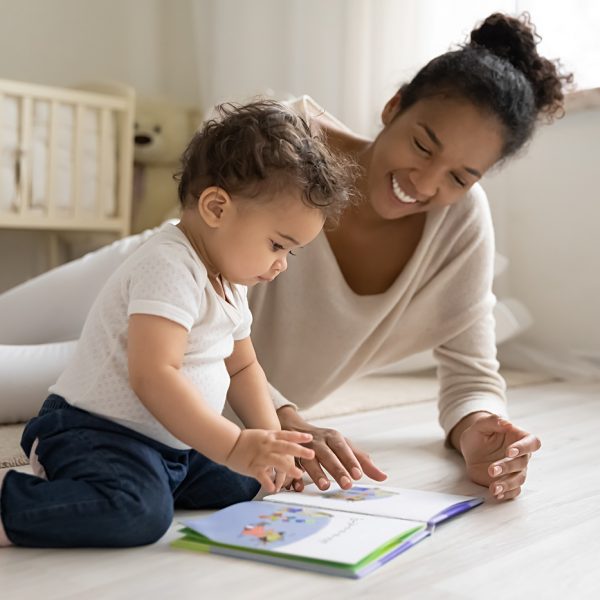 Smiling young african American mother sit on warm floor play with little infant toddler child, happy biracial mom relax have fun read book with small baby girl at home, motherhood, childcare concept